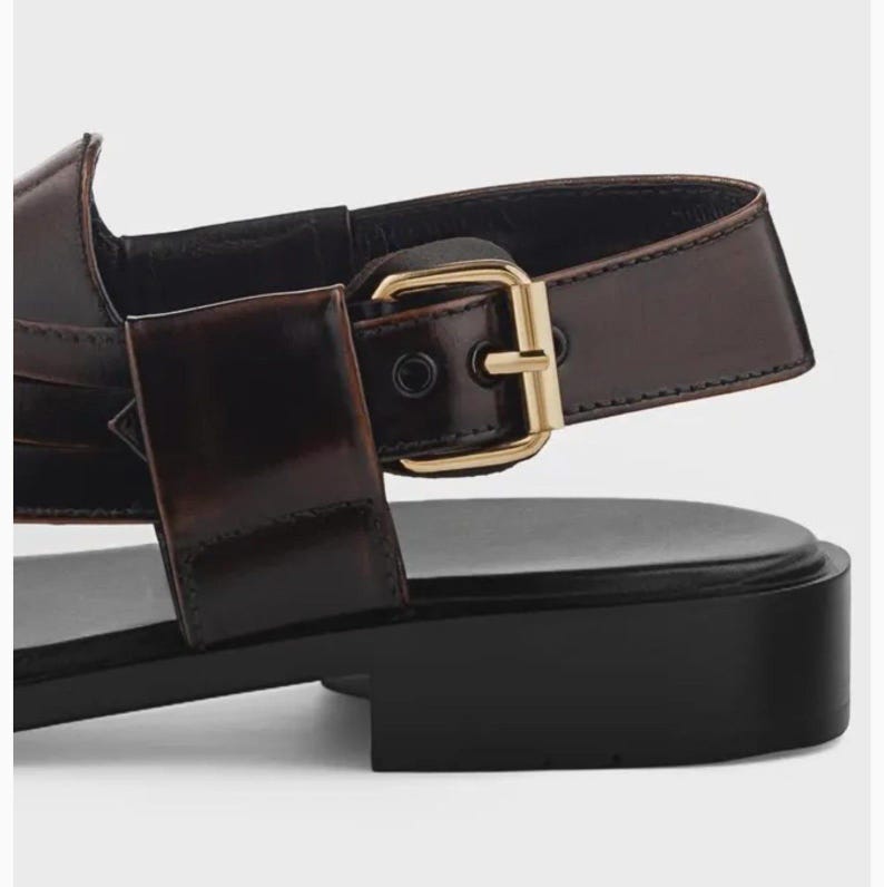K&ouml;nnte beinhalten: Eine dunkelbraune Ledersandale mit einer gl&auml;nzenden, goldfarbenen Metallschnalle am Seitenriemen. Das Schuhwerk verf&uuml;gt &uuml;ber eine glatte, flache schwarze Sohle und ein strukturiertes Fersenriemendesign mit einer gl&auml;nzenden Lederoberfl&auml;che.