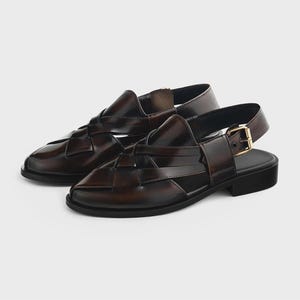 K&ouml;nnte beinhalten: Ein Paar dunkelbraune Ledersandalen mit einem geflochtenen Riemendesign am Vorderfu&szlig;, einer geschlossenen runden Zehenpartie und einem Fersenriemen mit goldfarbener Metallschnalle. Das Schuhwerk hat eine flache schwarze Sohle und einen Blockabsatz.
