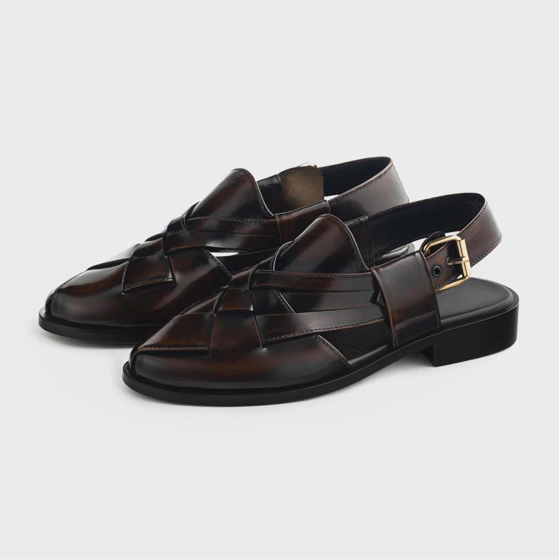 K&ouml;nnte beinhalten: Ein Paar dunkelbraune Ledersandalen mit einem geflochtenen Riemendesign am Vorderfu&szlig;, einer geschlossenen runden Zehenpartie und einem Fersenriemen mit goldfarbener Metallschnalle. Das Schuhwerk hat eine flache schwarze Sohle und einen Blockabsatz.