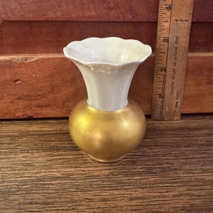 May include: A small vase with a bulbous gold-colored base and a flared, scalloped white rim. A wooden ruler placed next to the vase indicates a height of approximately 3.5 inches from the base to the top of the opening.