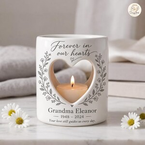 Könnte beinhalten: Ein weißer Kerzenhalter aus Keramik mit herzförmigem Ausschnitt, einem brennenden Teelicht und dem Text "Forever in our hearts", "Grandma Eleanor", "1948 - 2024" sowie "Your love still guides us every day" mit einem grauen Blumenranken-Design.