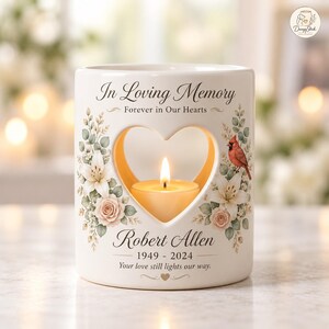 Könnte beinhalten: Ein weißer Kerzenhalter aus Keramik mit herzförmigem Ausschnitt, verziert mit Blumenillustrationen und einem roten Kardinal. Er zeigt den Text In Loving Memory, Forever in Our Hearts, Robert Allen, 1949-2024 und Your love still lights our way.