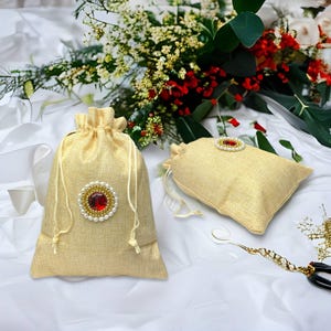 May include: Two beige burlap drawstring pouches featuring a circular embellishment on the front. Each ornament consists of a central red gemstone surrounded by a ring of small white pearls. The bags have matching beige fabric drawstrings.