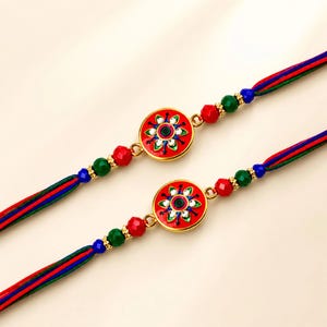 May include: Two decorative wristbands featuring a central circular gold-tone charm with red enamel and a floral pattern in green, blue, and white. The bands are made of multi-colored red, blue, and green threads, accented with faceted red, green, and blue beads.