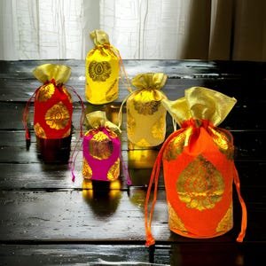 May include: Five drawstring pouches in red, yellow, gold, pink, and orange, each featuring a metallic gold floral pattern and a gold fabric top. The bags are arranged in a group, showcasing their vibrant colors and shiny, textured fabric construction.