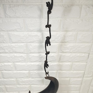May include: A dark brown, cast iron hanging planter featuring a vertical chain of three stylised monkeys linked together, suspended from a top hook. The bottom of the chain holds a crescent moon-shaped bowl designed to contain small items or plants.