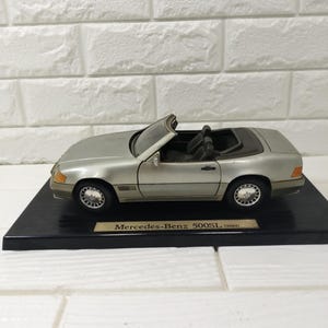 May include: A silver Mercedes-Benz 500SL (1989) scale model convertible car with a black interior, displayed on a black rectangular base with a gold-toned plaque featuring the text Mercedes-Benz 500SL (1989) centered on the front edge.