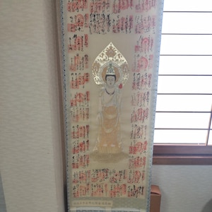 May include: A vertical hanging scroll featuring a central illustration of a standing figure with a gold halo, surrounded by columns of black Japanese calligraphy and numerous red square stamps on a cream-coloured fabric background with patterned borders.
