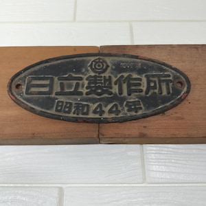 以下が含まれることがあります： 浮き彫りの文字とロゴが施された楕円形の黒い金属製プレート。日本語のテキストは「日立製作所」および「昭和４４年」と記されています。プレートの左右には取り付け用の穴が２つあり、黒い塗装の下から赤い塗装が見えるなど、経年による摩耗の跡が見られます。