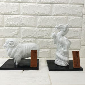 Puede incluir: Dos figuras de cerámica blanca, una con forma de carnero y otra de dragón, montadas sobre bases cuadradas negras. Junto a cada figura hay una pequeña placa de madera rectangular con caracteres de caligrafía china en color negro.