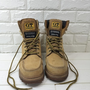 Puede incluir: Un par de botas de trabajo de cuero color canela con suelas de tacos marrones y cordones a rayas amarillas y negras. La lengüeta presenta un parche negro con el texto GT HAWKINS, GENUINE GOODYEAR WELT CONSTRUCTION e INSULATED en letras blancas.