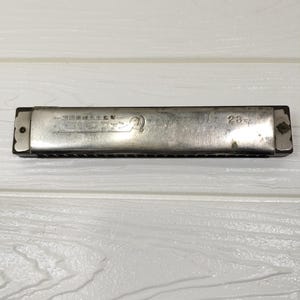 May include: A silver-toned metal harmonica with a dark brown body. The metal cover plate features engraved Japanese text, a circular logo, and the number 23. The instrument has visible screws on both ends and a series of rectangular air holes.