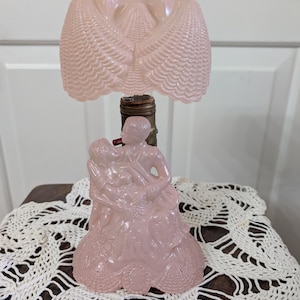May include: A pale pink glass table lamp featuring a sculpted base depicting two figures in an embrace. The lamp includes a matching pink glass shade with a textured, scalloped shell-like pattern and a metallic finial at the very top center.