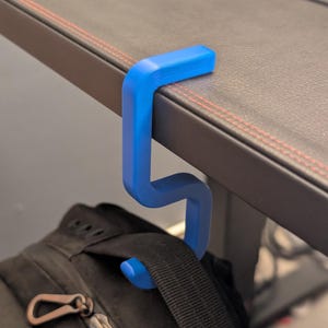 May include: A bright blue S-shaped desk hook designed to clamp onto the edge of a flat surface. The hook features a flat top section that rests on the desk and a curved lower section that supports the handle of a black backpack hanging below.