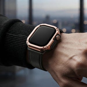 May include: A smartwatch featuring a metallic rose gold protective case and a dark grey leather strap. The watch face is black and rectangular with rounded corners, and the side includes a textured digital crown and a button for navigation.