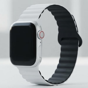 May include: A smartwatch with a silver rectangular case and a black screen. The watch features a side button and a digital crown with a red ring. It is equipped with a two-tone silicone band that is white on the exterior and black on the interior.