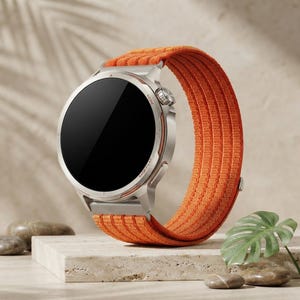 Puede incluir: Un reloj inteligente con caja metálica circular de color plateado y pantalla negra. El reloj cuenta con una correa de tela naranja texturizada con cierre de velcro. El lateral de la caja incluye una corona giratoria y un botón con marcas.