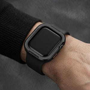 May include: A black smartwatch featuring a protective matte black bumper case surrounding the screen. The watch has a smooth black silicone band and a small orange accent button on the side, providing a sleek and functional look for the wrist.