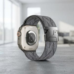 May include: A smartwatch with a metallic silver casing and a textured grey fabric strap. The strap features a black metal buckle mechanism. The back of the watch displays a circular sensor array and a side button with an orange accent ring.