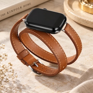 May include: A black smartwatch with a rectangular screen and a long, tan leather double-wrap watch band. The strap features white contrast stitching along the edges and a silver-toned metal buckle, designed to wrap twice around the wrist.