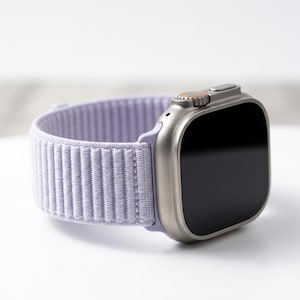 May include: A smartwatch with a metallic silver rectangular case and a black screen. The watch features a textured, light purple fabric strap with a ribbed design and a small orange accent on the side dial button of the watch casing.