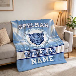 May include: A light blue and white fleece throw blanket featuring the text SPELMAN at the top and bottom, with the word NAME below it. The center displays a stylized jaguar head graphic enclosed within a circular Greek key border pattern.