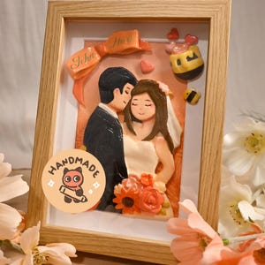 May include: A framed 3D clay art piece featuring a bride and groom in wedding attire, holding a bouquet of orange and pink flowers. An orange ribbon banner above reads John and Mary, accompanied by a small honey pot and bee figurine.