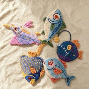 Può includere: Cinque toppe in tessuto ricamato con vari disegni di pesci. La collezione comprende un pesce spada rosa e blu, un pesce blu e crema, una rana pescatrice blu scuro con un'esca, un pesce a righe blu e giallo e un pesce azzurro.