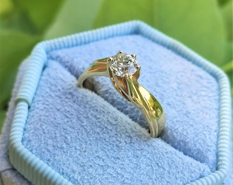 May include: A gold engagement ring with a single round diamond set in a twisted band. The ring is displayed in a blue velvet ring box.