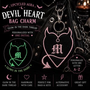 May include: A black heart-shaped bag charm with pink embroidered devil horns and a tail. The charm features the letter M in the center and includes a silver metal lobster clasp. The embroidery is made with glow-in-the-dark thread. Text includes UPCYCLED AURA DEVIL HEART BAG CHARM, GLOW IN THE DARK THREAD, PERSONALIZED WITH ONE INITIAL, and PERSONALIZE WITH ONE LETTER A-Z.