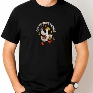 Könnte beinhalten: Ein schwarzes Kurzarm-T-Shirt mit einer Grafik einer weißen Gans, die einen gelben Schutzhelm trägt und einen Werkzeuggürtel sowie ein Stromkabel hält. Der Text "OFF TO WIRE THINGS" ist in einem Bogen mit gelben Blitzen darüber gedruckt.