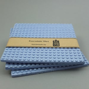 Può includere: Un set di quattro piastrelle in porcellana blu chiaro con un'etichetta marrone che recita "Porcelain Tiles Handmade by [logo]"