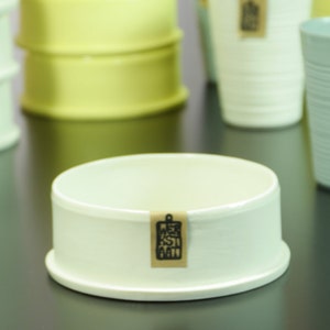 May include: A white ceramic pedestal with a brown label that says "KESTI ART". The pedestal is on a black surface with other ceramic bowls and cups in various colors.
