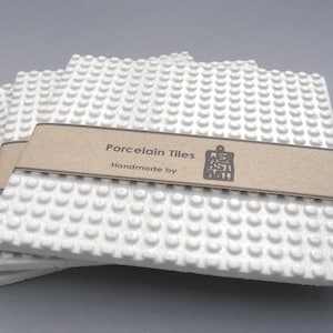 Puede incluir: Tres azulejos de porcelana blancos con un patrón de puntos en relieve. Los azulejos están apilados uno encima del otro y tienen una etiqueta de papel marrón que dice "Porcelain Tiles Handmade by Mesa Arts".