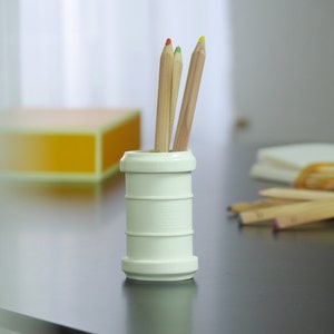 May include: A white ceramic pencil holder with three wooden pencils with coloured tips. The pencils are red, green, and yellow.