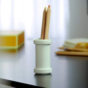 May include: A white ceramic pencil holder with three wooden pencils inside. The pencils are yellow, orange, and red. The holder is on a black table.