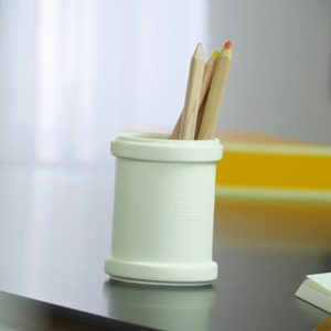 Puede incluir: Un porta lápices de plástico blanco con cuatro lápices de colores dentro. Los lápices son rojo, naranja, amarillo y verde. El porta lápices está sobre una mesa de madera.