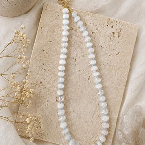 May include: A beaded necklace featuring alternating translucent white and pale blue circular beads. The necklace is finished with a gold-toned metal lobster claw clasp and a short extender chain, providing a secure closure for the jewelry piece.