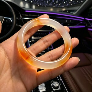 Może przedstawiać: Półprzezroczysta, okrągła bransoletka typu bangle wykonana z gładkiego, polerowanego kamienia. Materiał ma mlecznobiały kolor bazowy z wyraźnymi, żywymi smugami w odcieniach głębokiego pomarańczu i czerwonawego brązu, które zdają się świecić pod światło.
