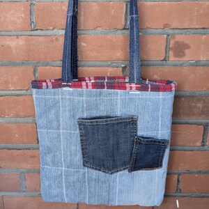 May include: A tote bag crafted from light blue denim with a white grid pattern, featuring two dark blue denim pockets on the front. The bag has a red and black plaid fabric trim along the top edge and two dark blue denim shoulder straps.