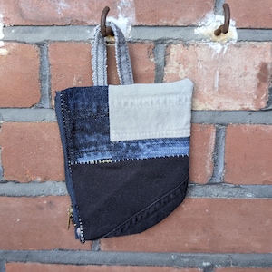 May include: A small zippered pouch constructed from patchwork fabric panels in shades of dark blue denim, light grey denim, and solid black fabric. The pouch features a grey fabric loop handle at the top and a brass-coloured metal zipper closure.