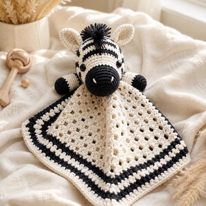 Pode incluir: Uma manta de segurança de crochê com uma cabeça de zebra acolchoada com listras pretas e brancas, crina preta e olhos de segurança pretos. A manta é feita de fio cor de creme com um padrão de ponto granny e uma borda preta e branca.
