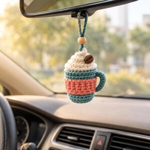 Può includere: Un ornamento all'uncinetto a forma di tazza di caffè appeso allo specchietto retrovisore. La tazza è color verde acqua con una fascia corallo, sormontata da panna montata bianca e un chicco di caffè marrone. Una perlina di legno è sul cordino.