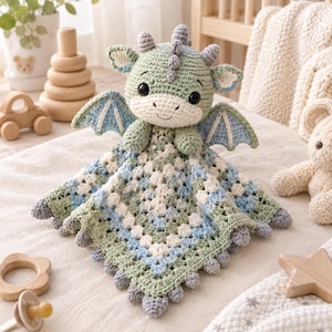May include: A crochet security blanket featuring a stuffed sage green dragon head with black eyes, small horns, and blue wings. The blanket body is a square granny square pattern in shades of sage green, light blue, and cream with small grey bobbles.