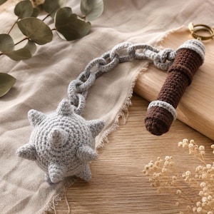 May include: A crocheted flail keychain featuring a grey spiked ball attached to a grey crocheted chain and a brown cylindrical handle with grey accents. The handle is topped with a metal keyring. The entire object is crafted from yarn.