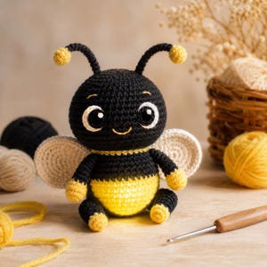 Puede incluir: Un juguete de abeja tejido a ganchillo con una cabeza redonda negra, grandes ojos blancos y negros, dos antenas con puntas amarillas y un cuerpo con rayas negras y amarillas. La abeja tiene pequeñas alas color crema y extremidades amarillas.
