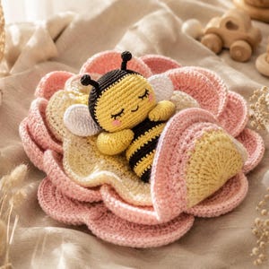 Puede incluir: Un juguete de abeja tejido a ganchillo con cuerpo a rayas amarillas y negras, cabeza negra y alas blancas, descansando dentro de una manta tejida en forma de flor de varias capas en rosa suave y amarillo pálido con bordes festoneados.