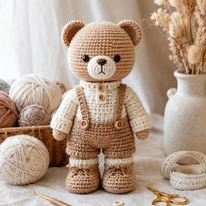 Könnte beinhalten: Ein gehäkelter Teddybär mit hellbraunem Fell, bekleidet mit einem weißen langärmeligen Hemd und einer braunen Latzhose mit Knöpfen. Der Bär hat schwarze Augen, eine braune Nase und gestickte Munddetails. Er trägt dazu passende braune Schuhe.