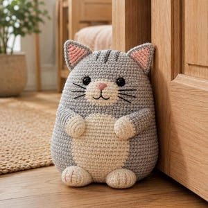 Puede incluir: Un tope de puerta de ganchillo con forma de gato en color gris y crema, con cuerpo redondeado, orejas puntiagudas con detalles rosados, ojos de plástico negro, nariz bordada en rosa y bigotes. Tiene pequeñas patas delanteras y traseras.