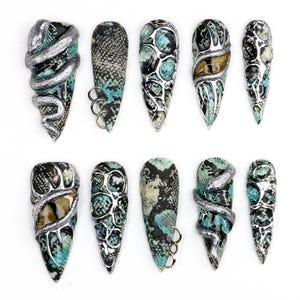 May include: A set of ten press-on nails featuring a turquoise and black snakeskin pattern. The designs include silver metallic snake accents, three-dimensional circular rings, textured silver overlays, and realistic eye motifs with gold and black irises.